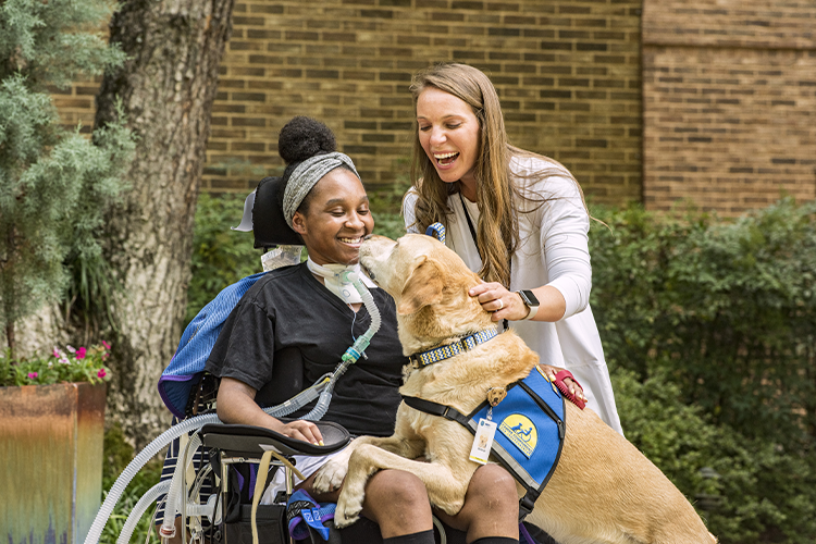 Patient outside on ventilator in Shepherd Center's Secret Garden with her respiratory therapist. A facility dog jumps up into her lap to giver her a kiss.